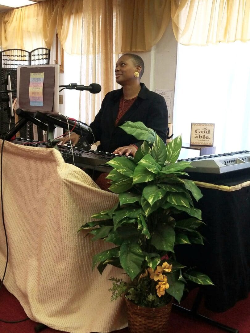 A person standing at a lectern with a microphone and audio equipment, with a plant in the foreground.