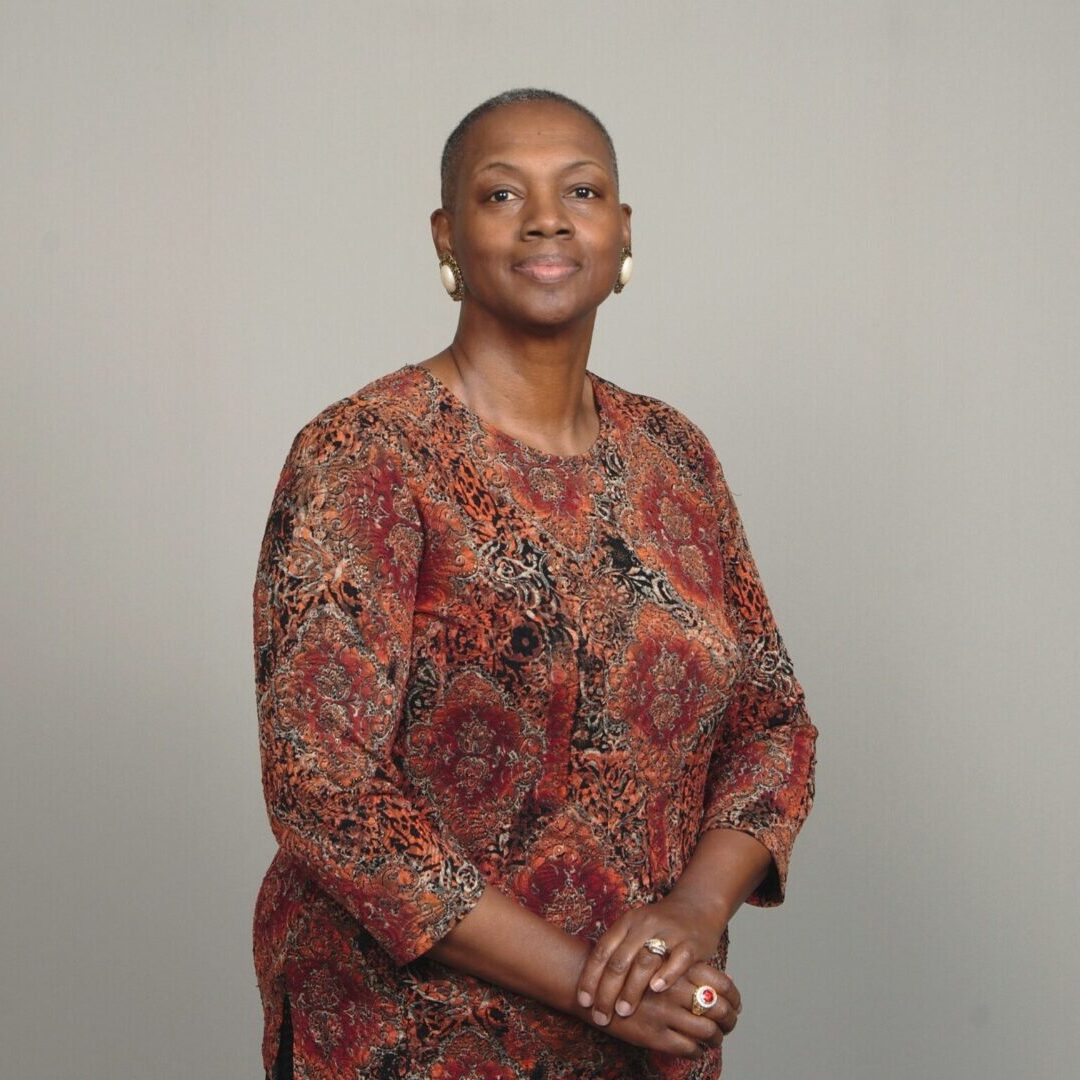 A woman with short hair wearing a patterned top smiles while posing with her arms crossed.